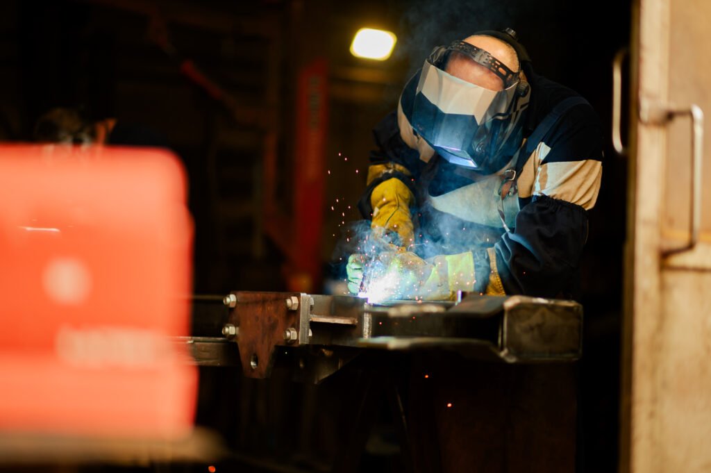 Male welder wearing construction gloves and welding mask hardworking and welding with welding machine metal in workshop. Worker in furniture factory grinding metal.