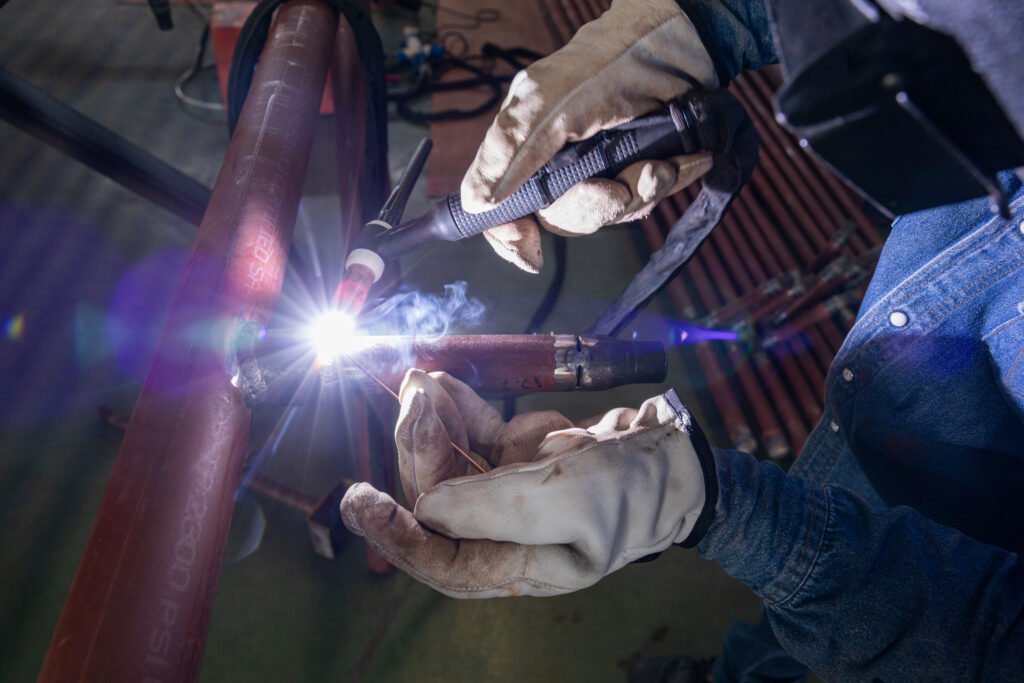 A skilled welder using TIG welding on a metal pipe, with protective gloves and helmet, producing bright light and precision work in an industrial setting.